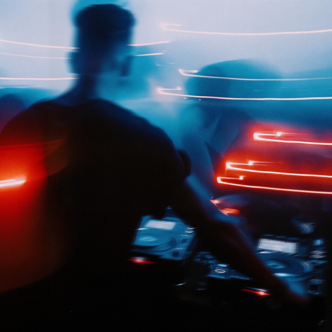 Blurred photo of a DJ performing in a dark, neon-lit nightclub with streaks of red and blue light creating a dynamic, energetic atmosphere.