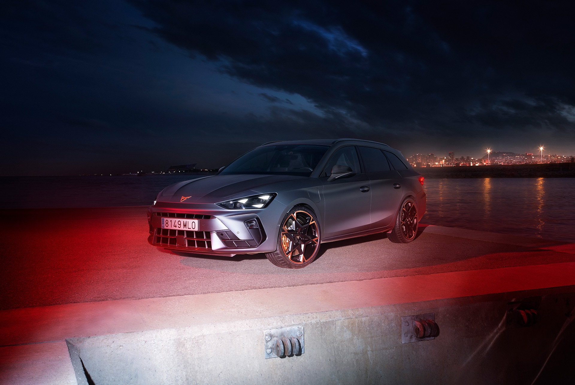 A CUPRA Leon Sportstourer 2025 in matte grey is parked on a waterfront at night. The city skyline is visible in the background, with its lights reflecting in the water. The car is illuminated by red lights, creating a dramatic and eye-catching scene.