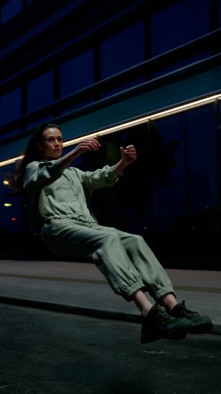 A woman in a light green jumpsuit and dark sneakers appearing to float in a seated position on a city street at night, with a modern glass building in the background.